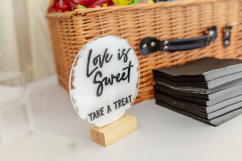 Acrylic 'Love is Sweet, Take a Treat' sign on wood stand beside black napkins and wicker basket.