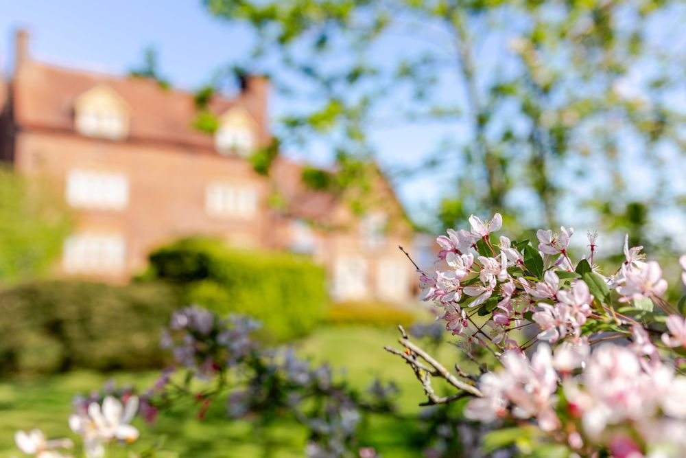 Pink spring blossoms in foreground with a blurred red brick country house and lush green garden in background.