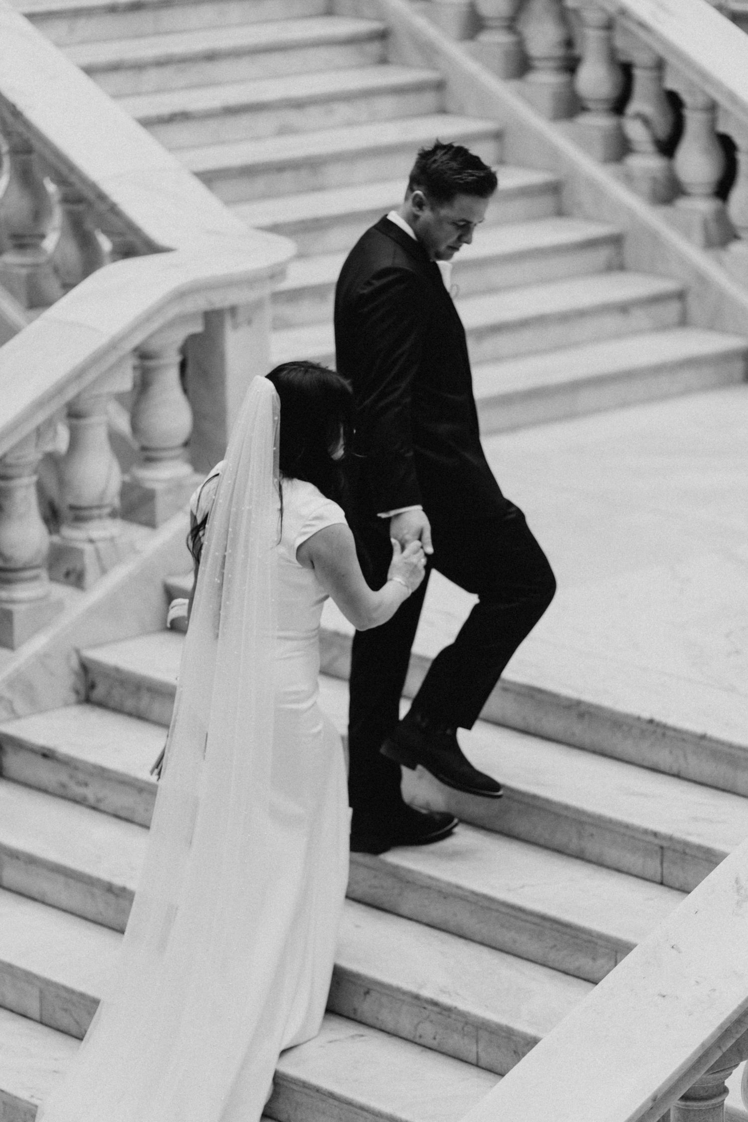 Black and white photo of bride and groom holding hands walking up marble stairs on their wedding day.