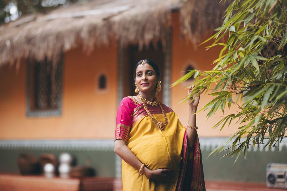 Pregnant woman in yellow and pink saree with gold jewelry standing outdoors near bamboo by a rustic thatched building.