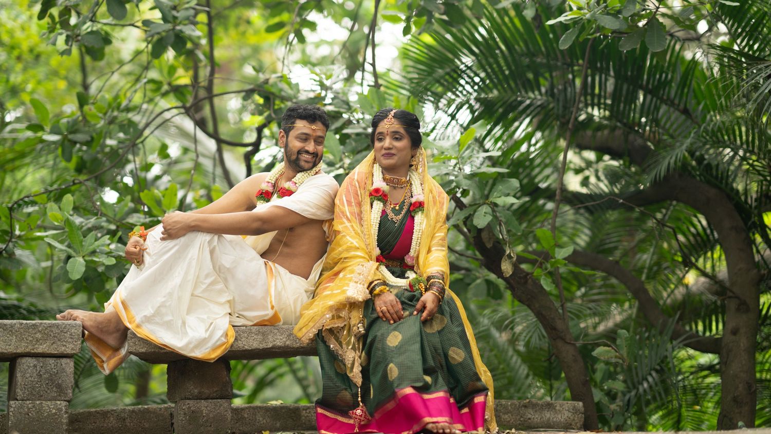 Indian couple in traditional attire smiling outdoors, man in white dhoti and woman in green saree, lush greenery background.