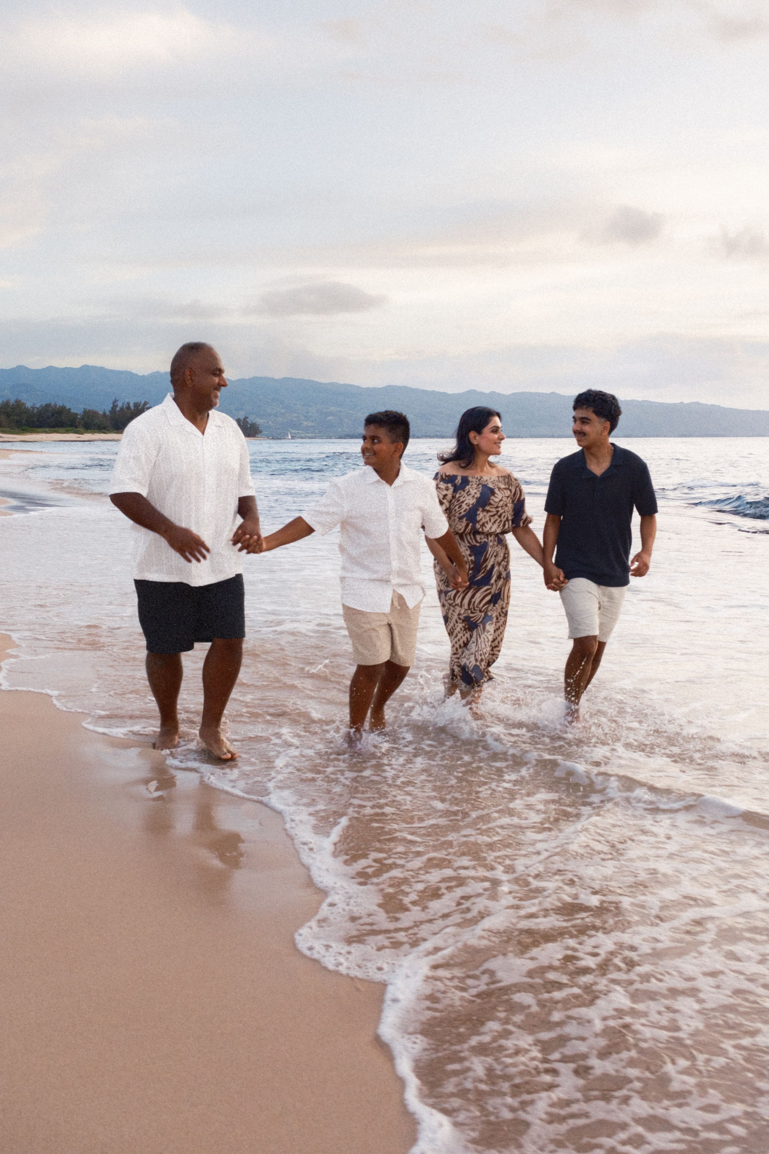 Family of four walking along sandy beach shoreline at sunset, waves washing over their feet.