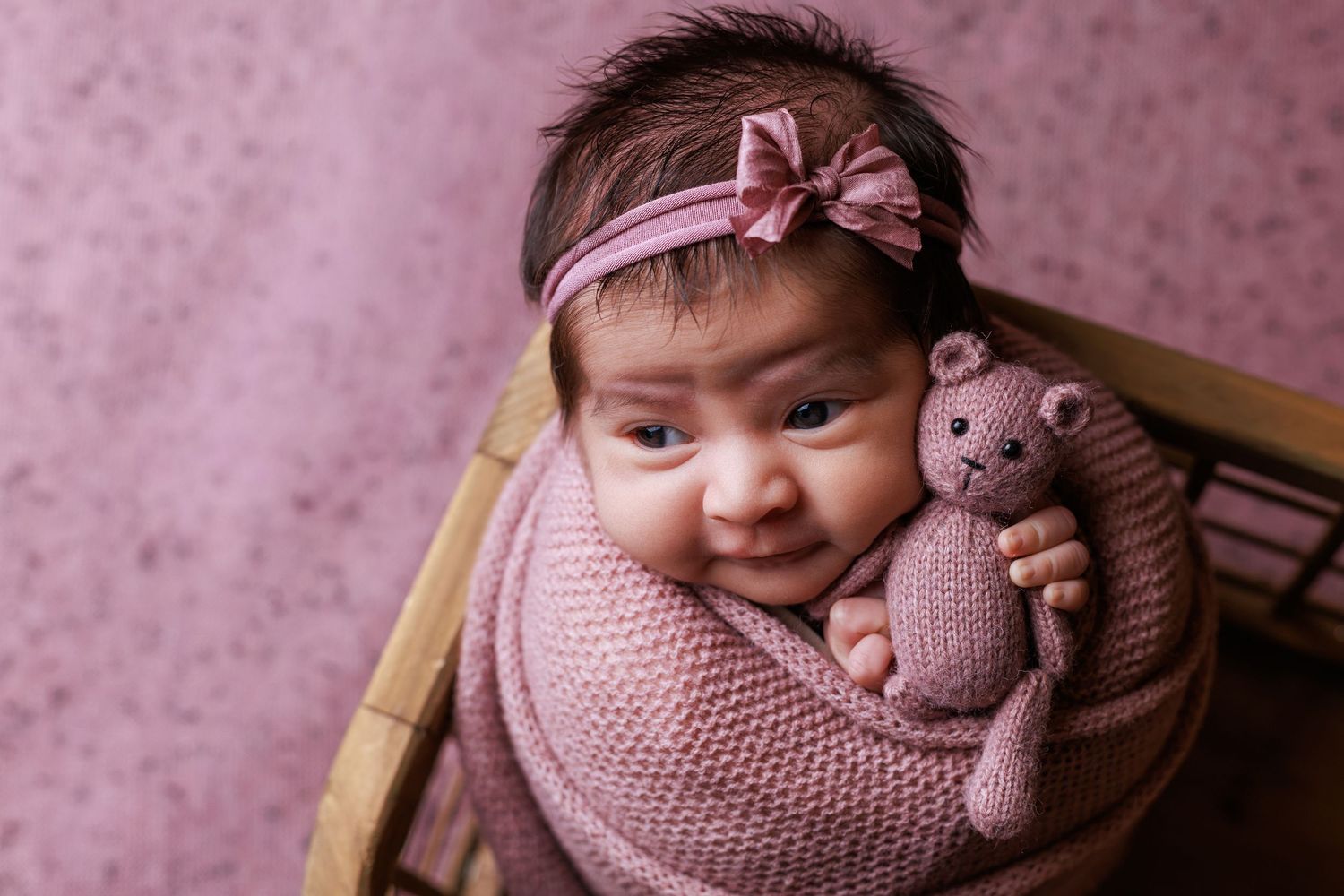 Newborn baby girl wrapped in pink knit swaddle holding teddy bear, wearing pink bow headband in wooden basket.