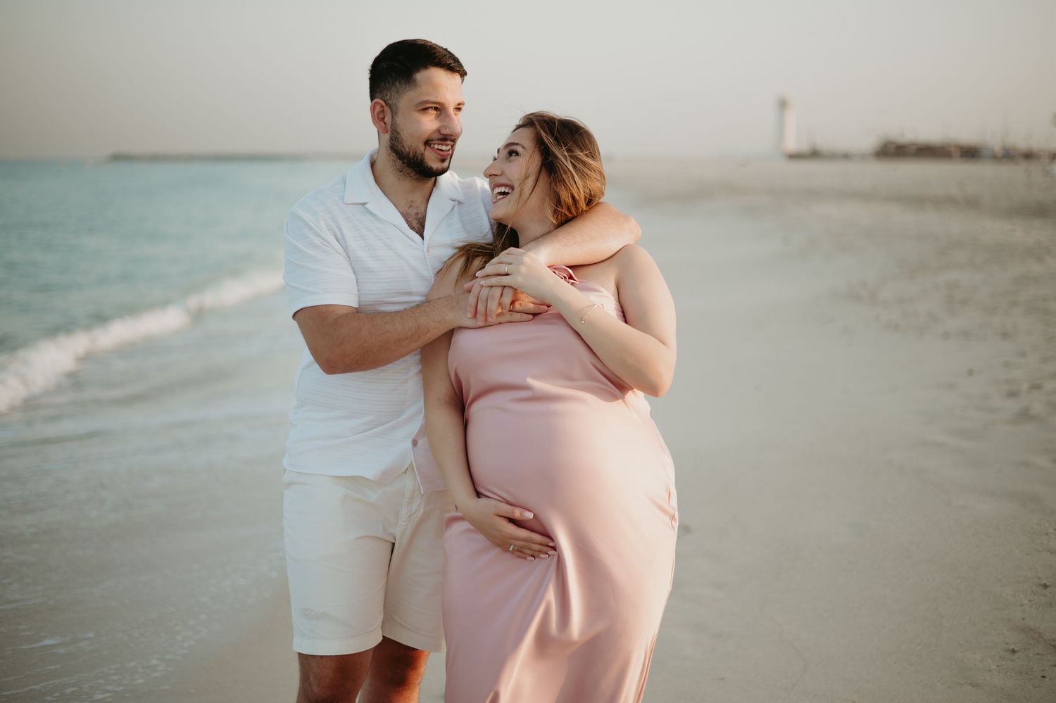 Couple sharing a tender moment on a sandy beach, woman in pink maternity dress cradling her baby bump at sunset.