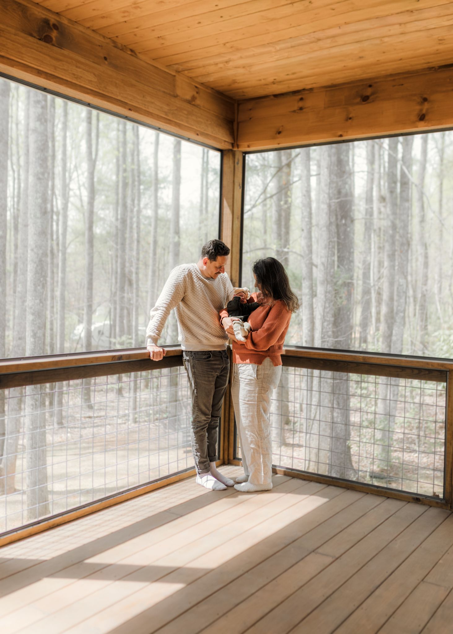 A couple stands on a screened wooden deck in a forest cabin, tenderly looking at their newborn baby.