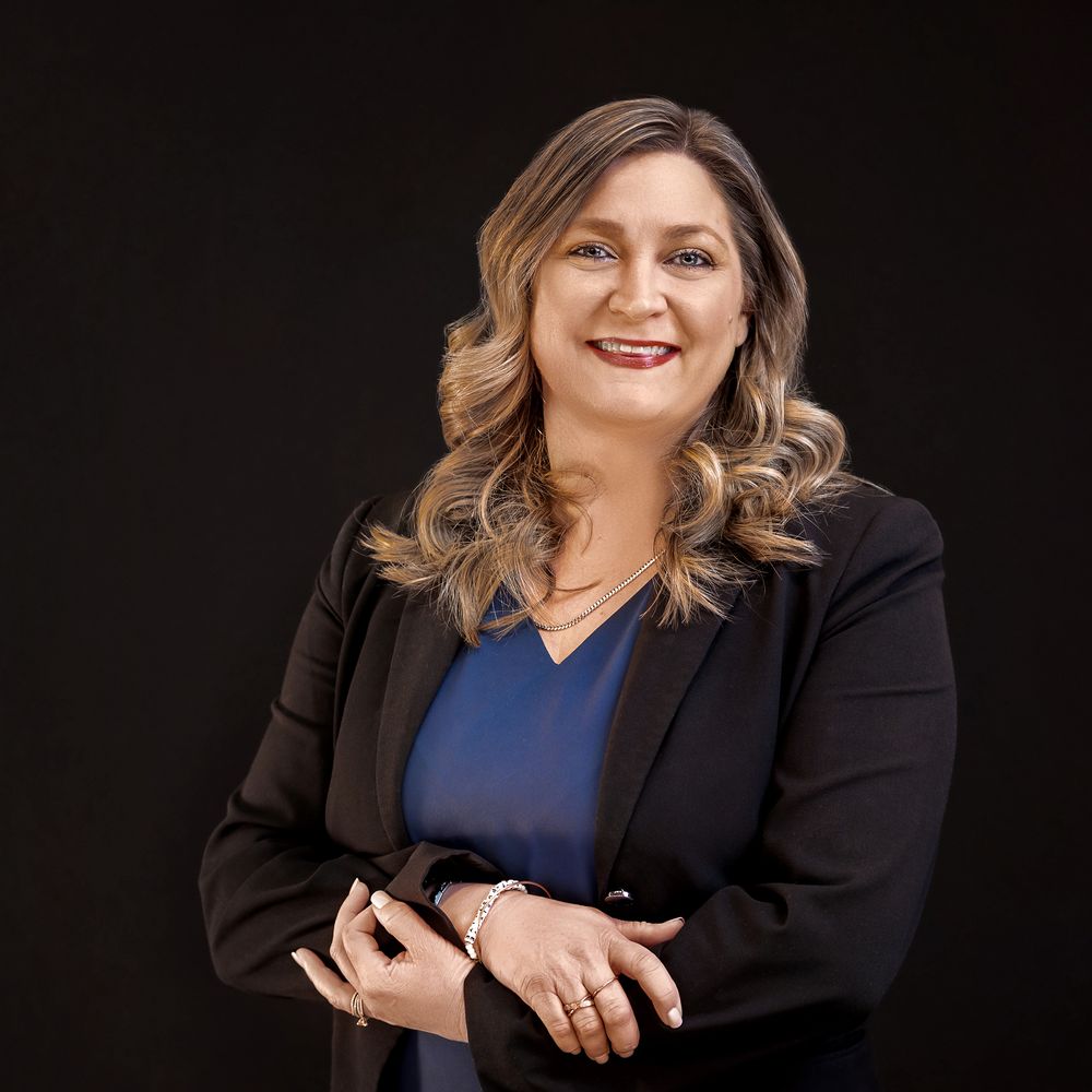 Professional woman in black blazer and blue top smiling confidently against dark background.