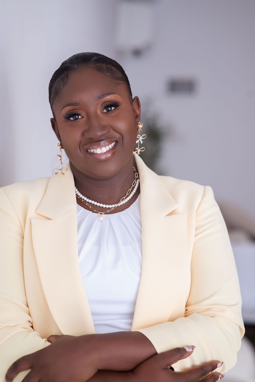 Smiling woman in yellow blazer and white top, wearing gold earrings and pearl necklace, arms crossed, professional portrait.