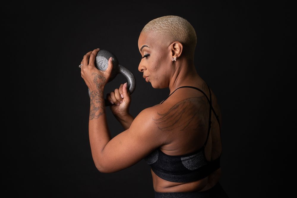 Muscular woman with shaved blonde hair lifting a kettlebell in a black sports bra against a dark background.