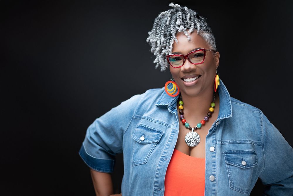 Smiling woman with gray locs, colorful necklace, red glasses, and denim jacket poses confidently against dark background.
