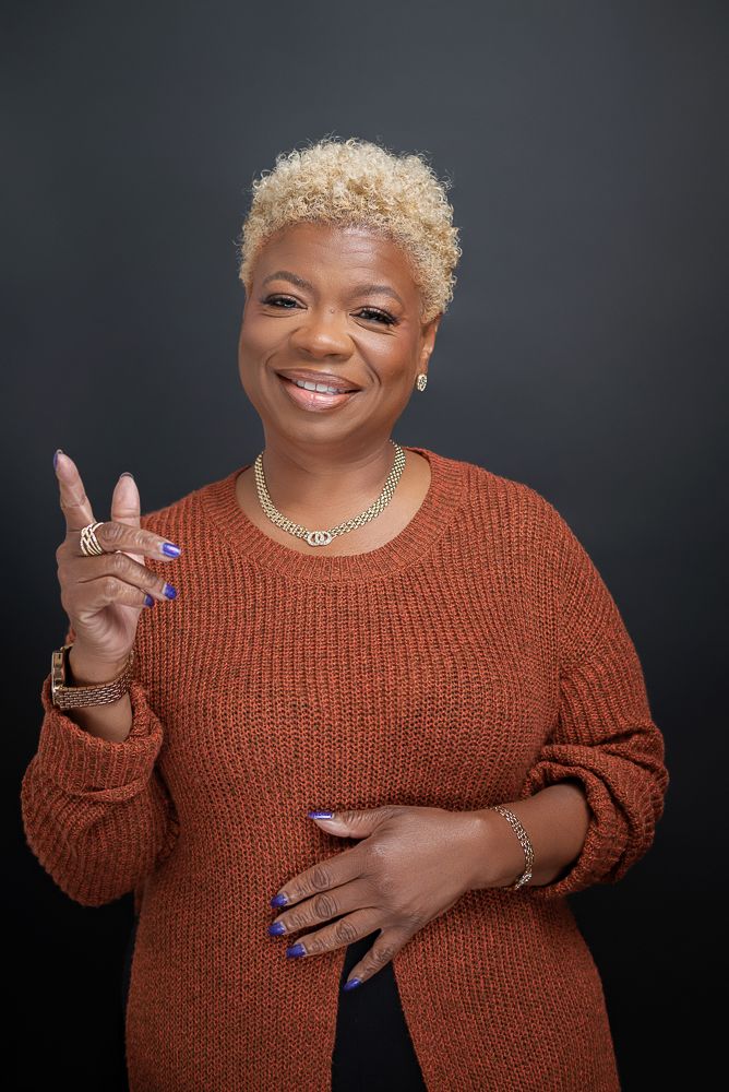 Smiling woman with blonde curly hair in rust sweater and gold jewelry, pointing upward against dark background.