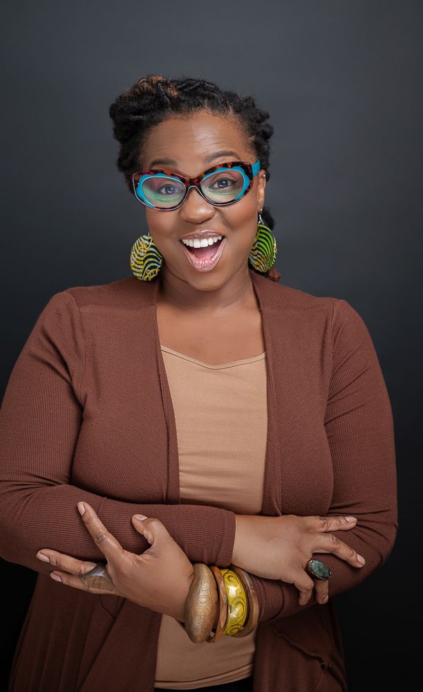 Smiling woman with colorful cat-eye glasses, yellow earrings, brown cardigan, and gold bracelets posing confidently.