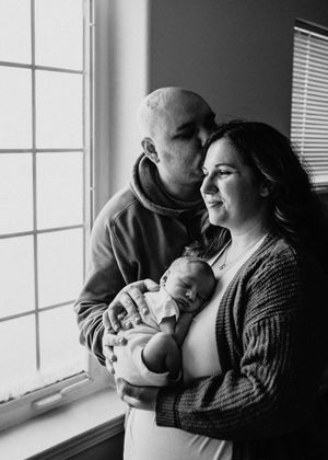 Black and white portrait of parents holding their newborn baby together near a window during an in-home newborn session