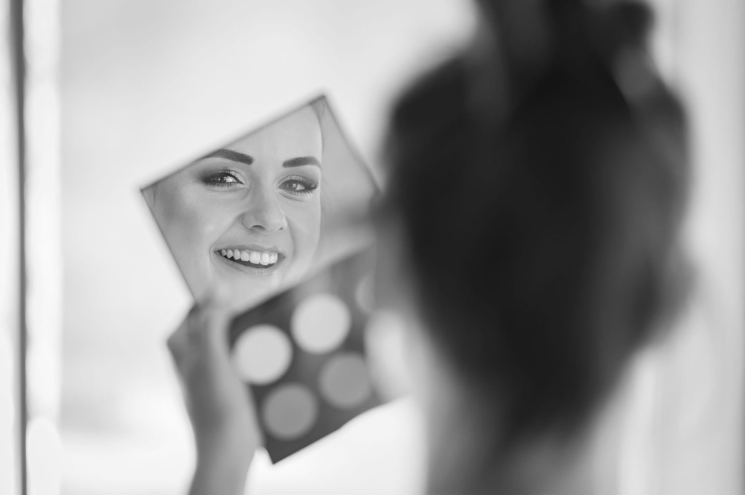 Black and white photo of a smiling bride checking her makeup in a compact mirror while getting ready.