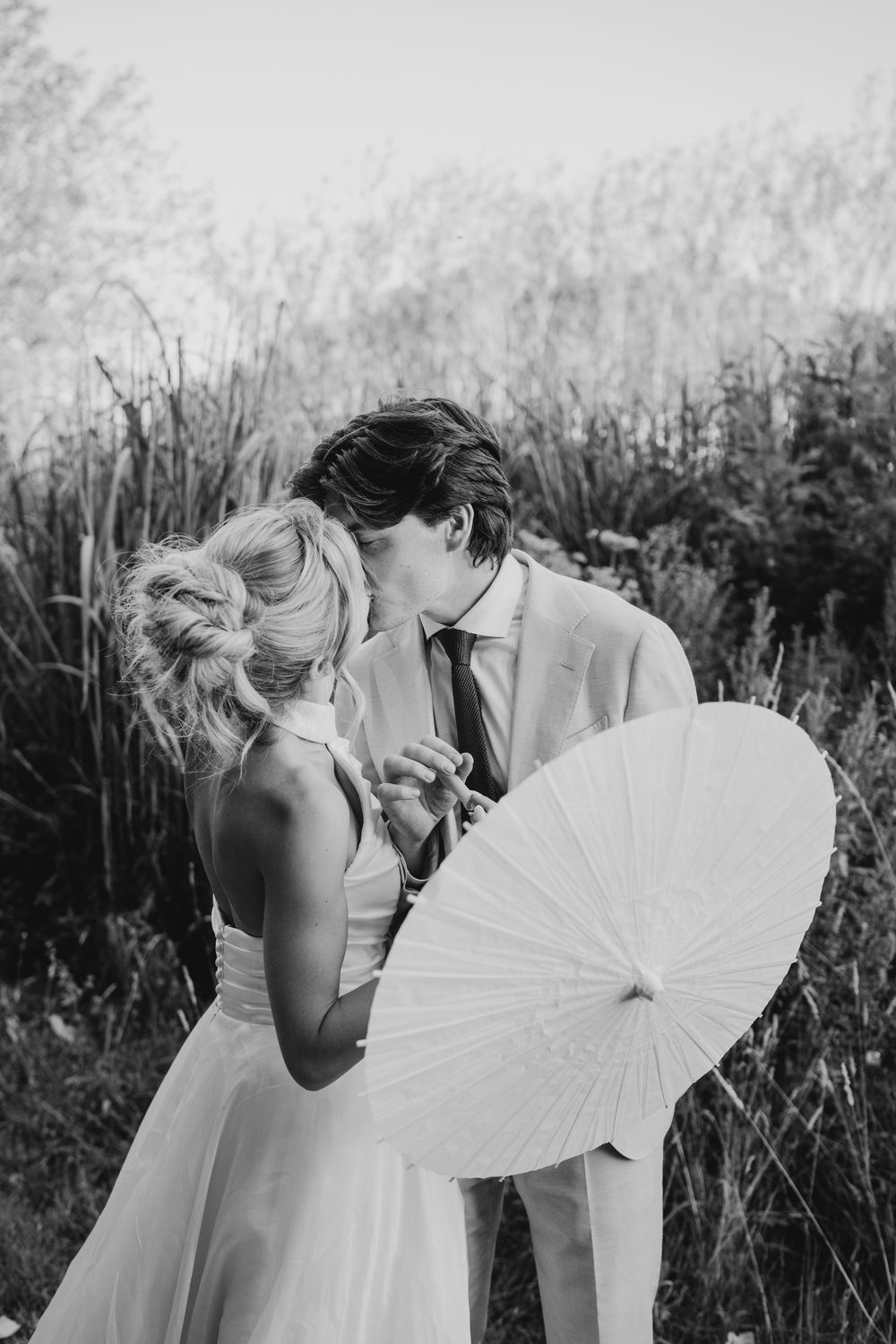 Black and white photo of a bride and groom sharing a kiss while holding a white parasol umbrella outdoors.