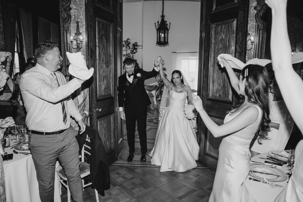 Black and white photo of a bride and groom entering their wedding reception while guests celebrate with raised hands.