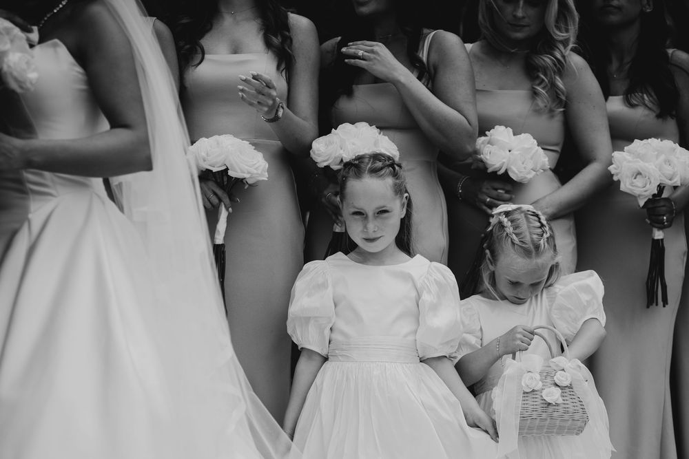 Black and white photo of flower girls in white dresses with floral crowns standing among bridesmaids at a wedding.
