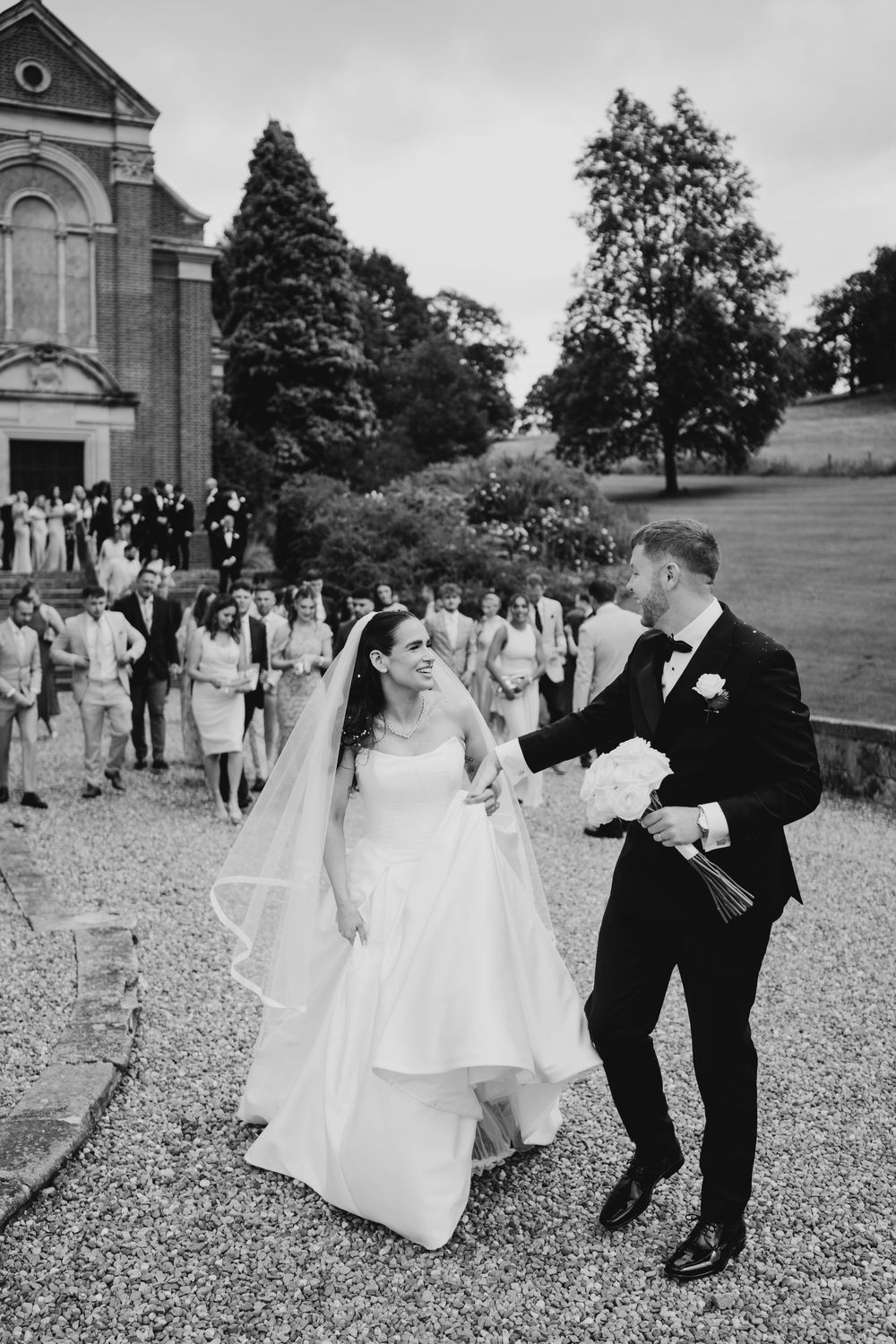 Black and white photo of bride and groom walking together after wedding ceremony with guests in background.