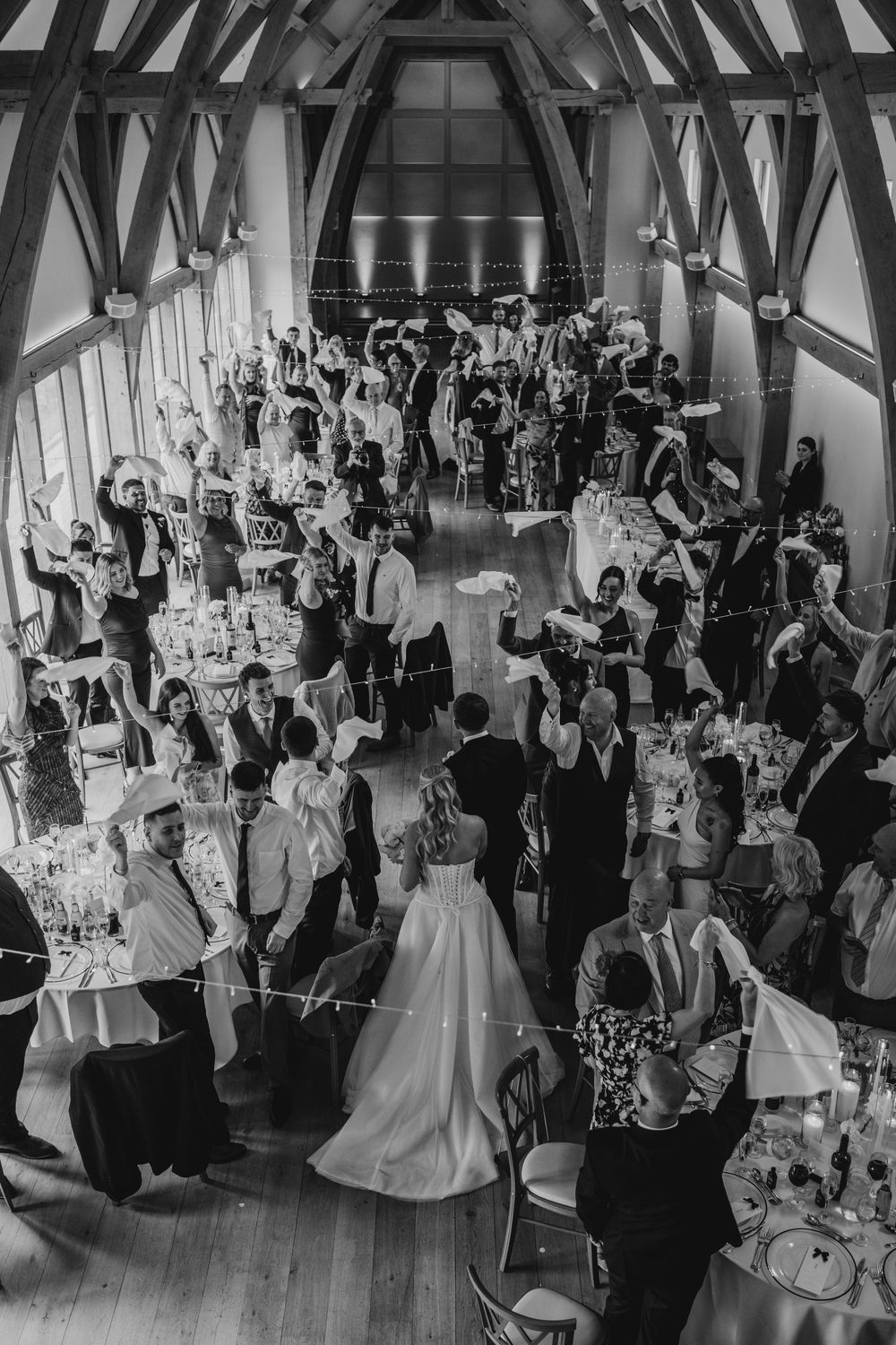 Black and white aerial view of a wedding reception in a barn venue with guests raising hands in celebration.