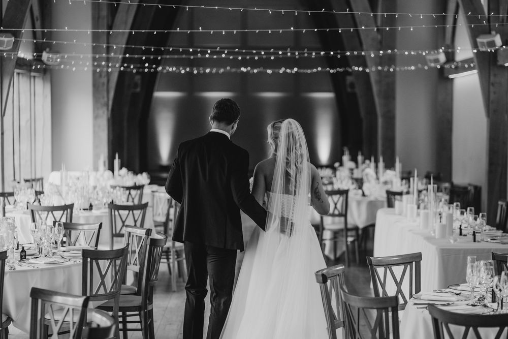 Black and white photo of bride and groom walking into elegant wedding reception hall with fairy lights.