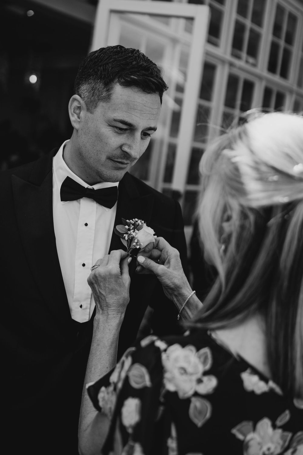 A woman pins a boutonniere on a man in a tuxedo at a wedding in a black and white photo.