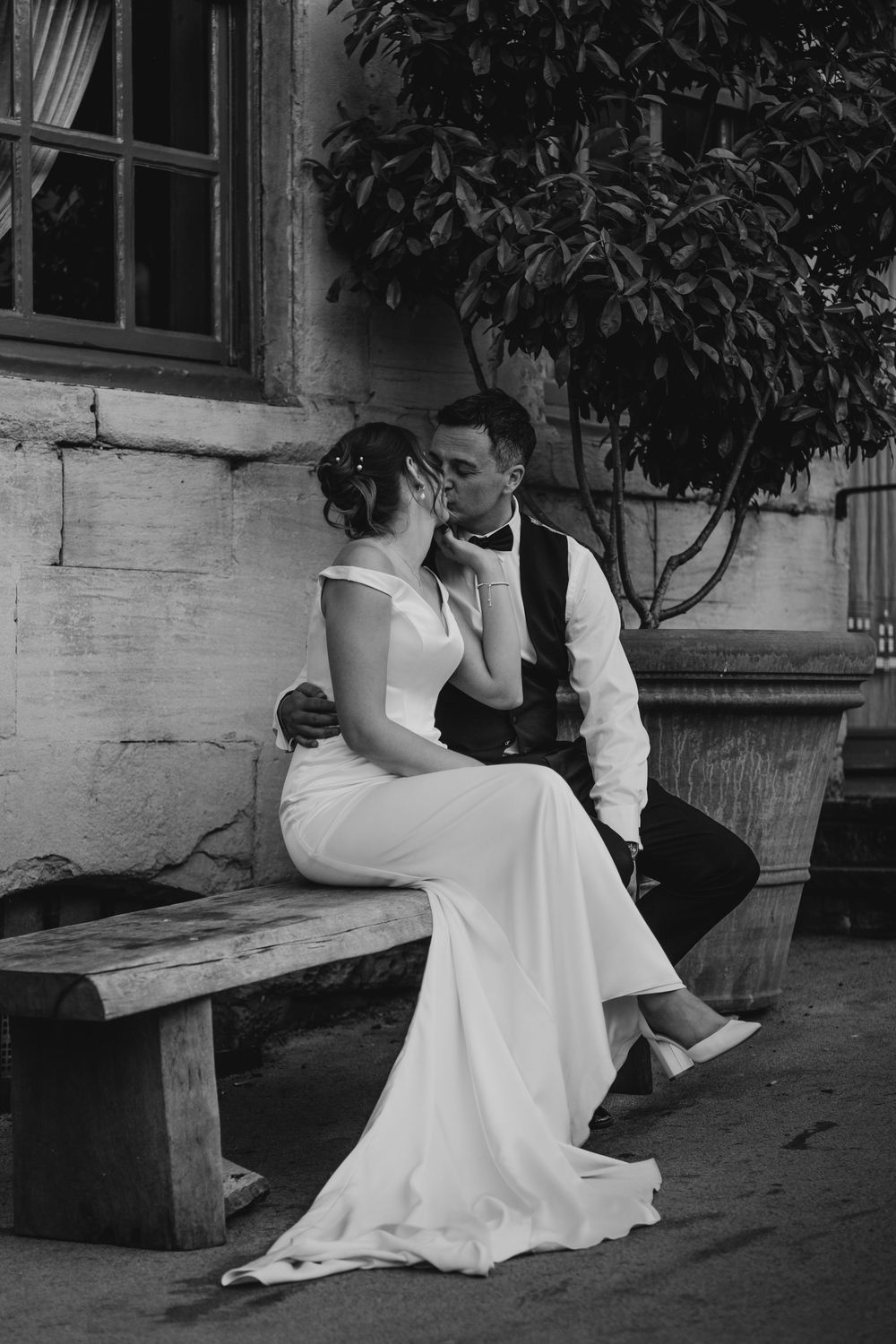 Black and white photo of a bride and groom sharing a kiss while seated on a wooden bench outside a rustic building.