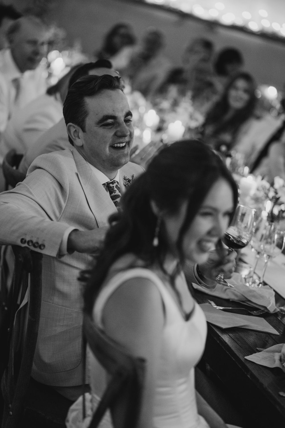 Black and white wedding photo of smiling groom in suit beside bride holding wine glass at reception dinner.