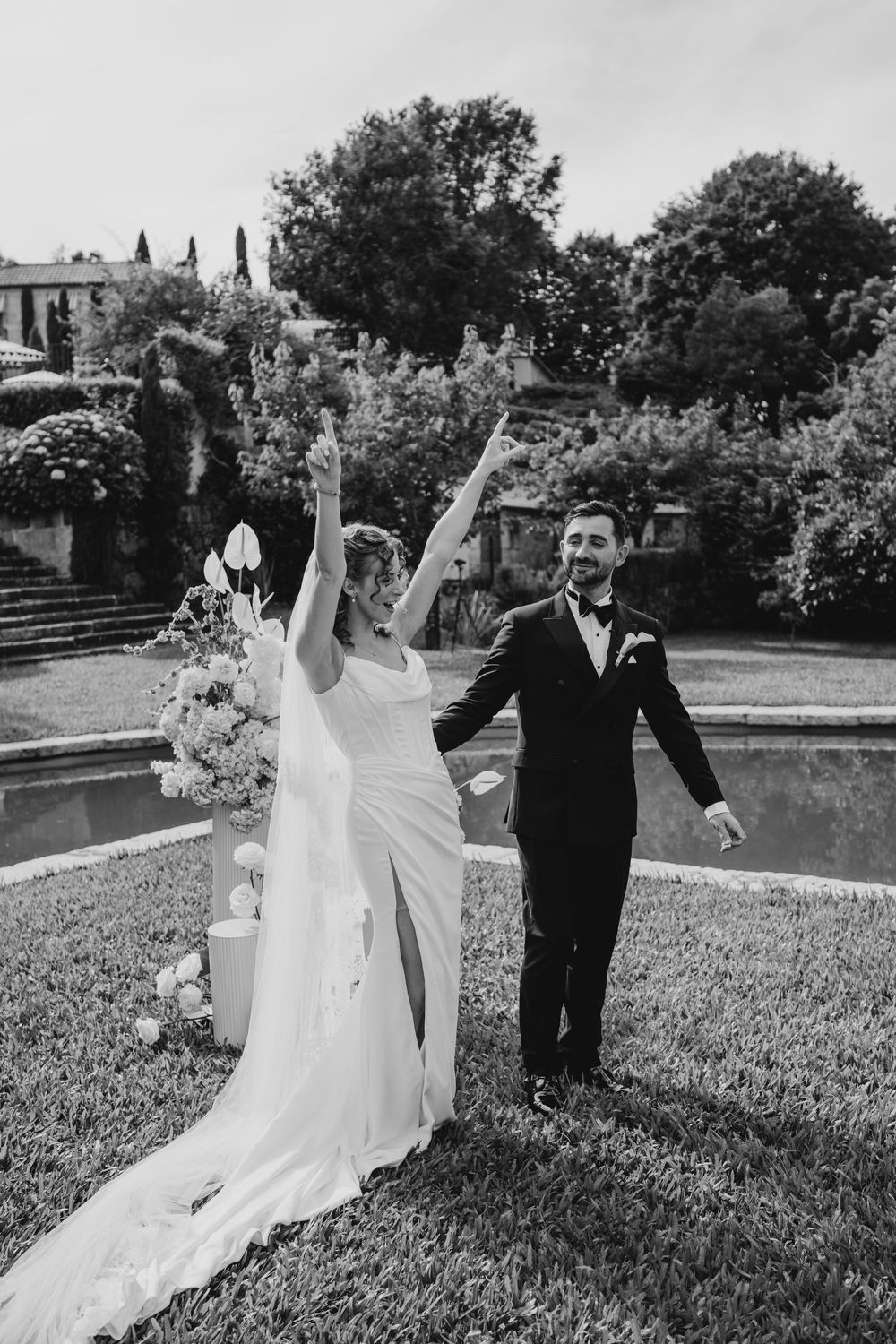 Joyful bride and groom celebrate outdoors at elegant garden wedding venue, raising arms in triumph near ornamental pond.