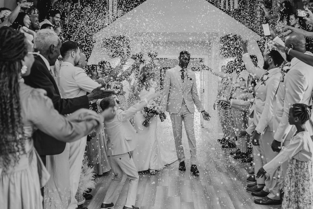 Black and white wedding photo of couple walking down aisle through confetti shower with cheering guests.