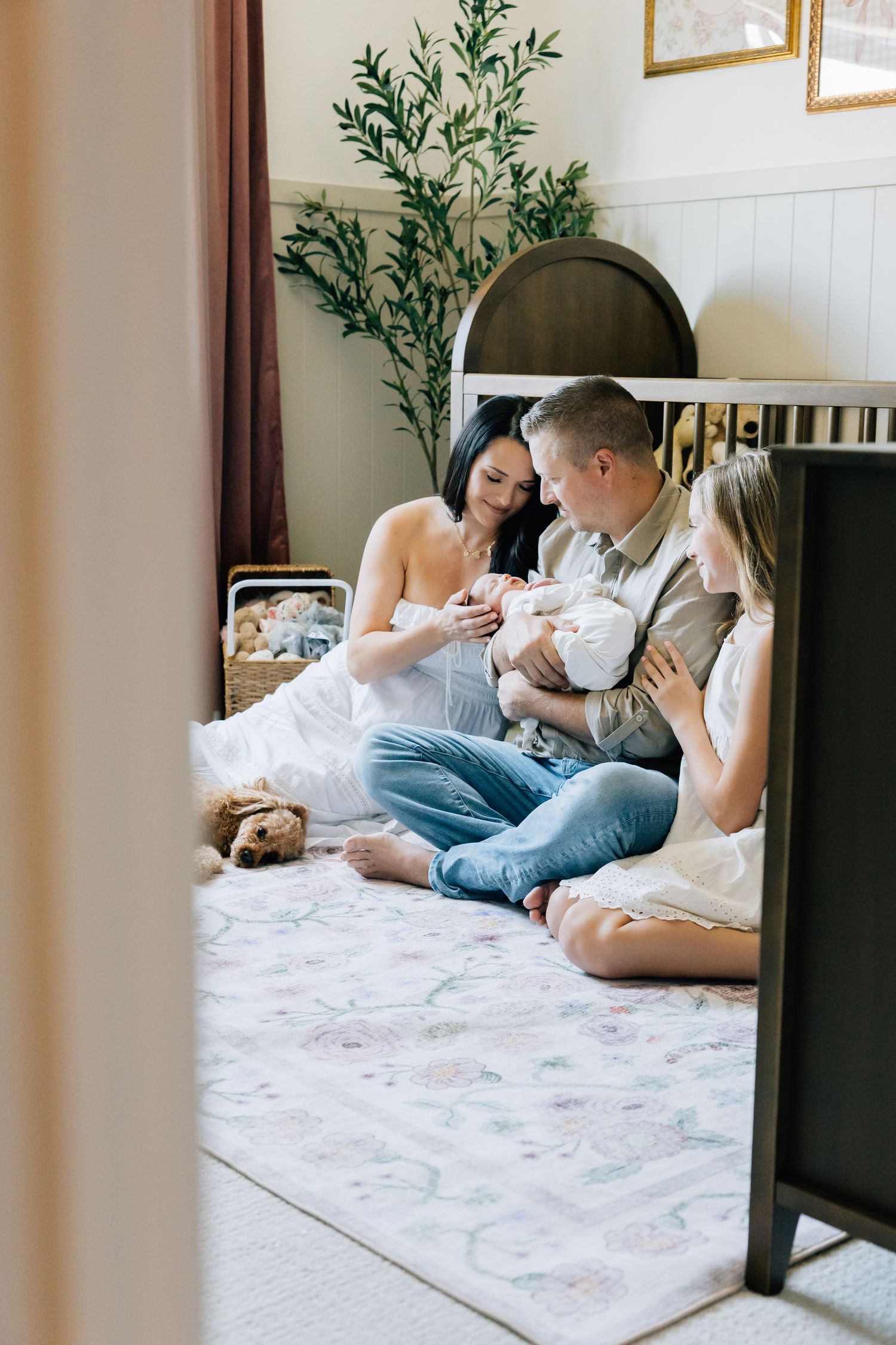 Family sitting on nursery floor admiring their newborn baby, with an older sibling joining the tender moment.