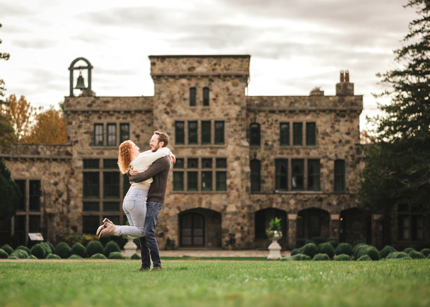 Engaged couple laughing and embracing on the lawn in front of the historic stone Ames Mansion at Borderland State Park.