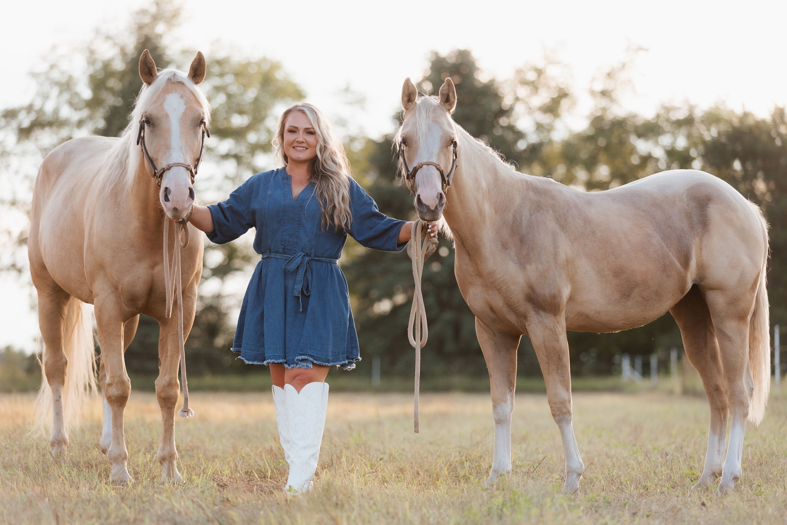 Woman in blue dress and white boots stands between two palomino horses in a sunny field.