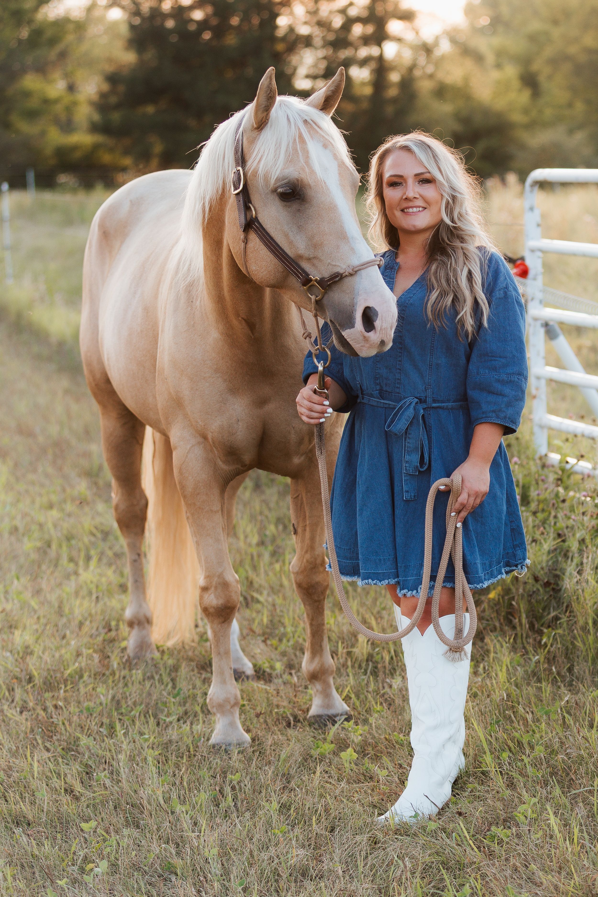 Smiling woman in denim dress and white boots stands beside a palomino horse in a sunny field near a metal gate.
