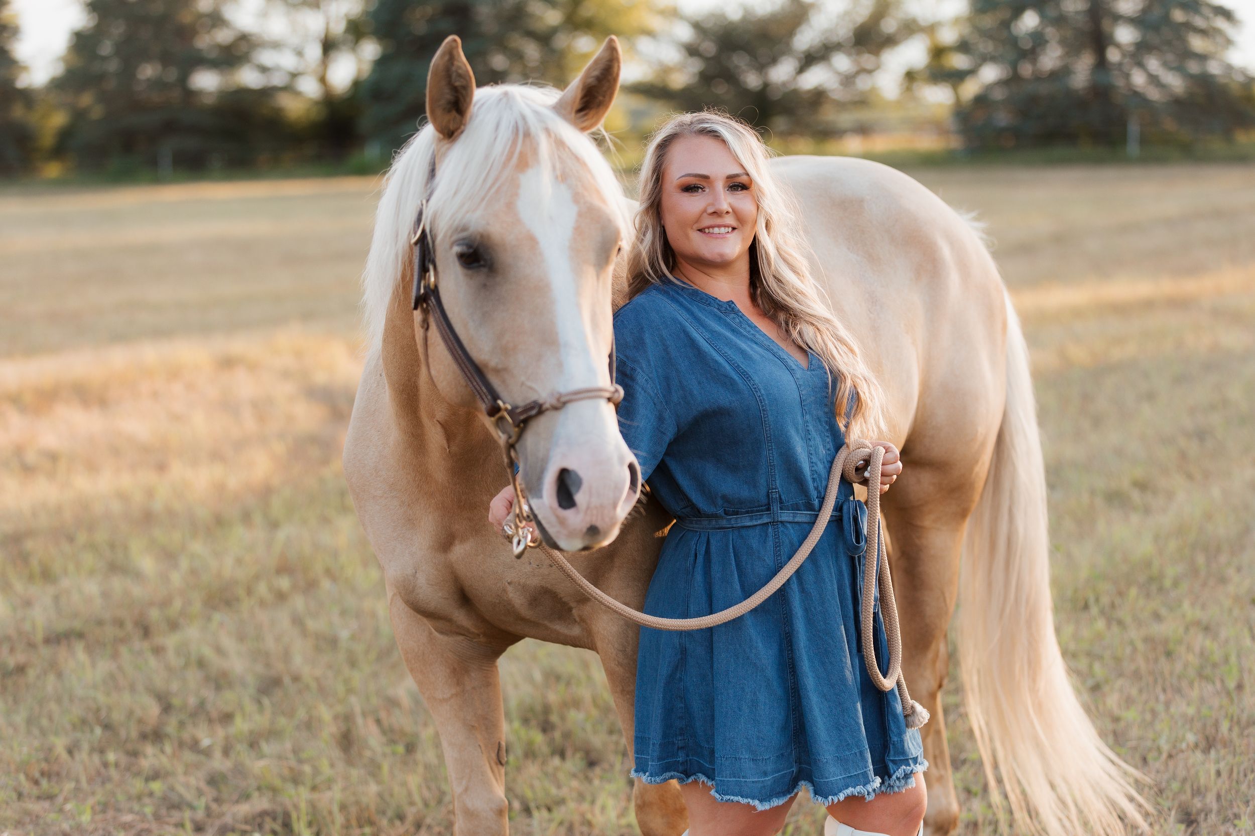 Young blonde woman in blue dress poses with a palomino horse in a golden sunlit field.