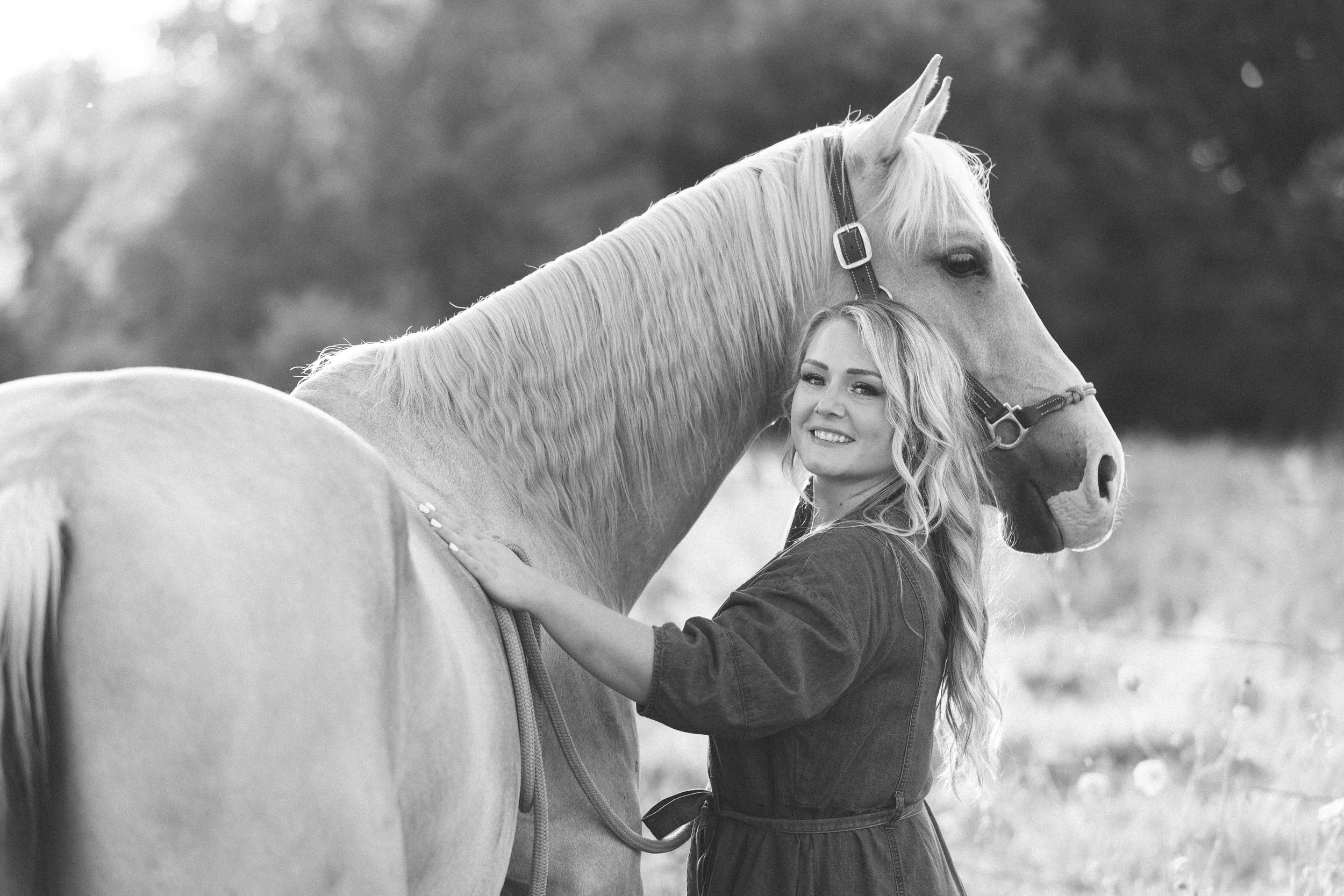 Black and white photo of a smiling blonde woman embracing a light-colored horse in a field.