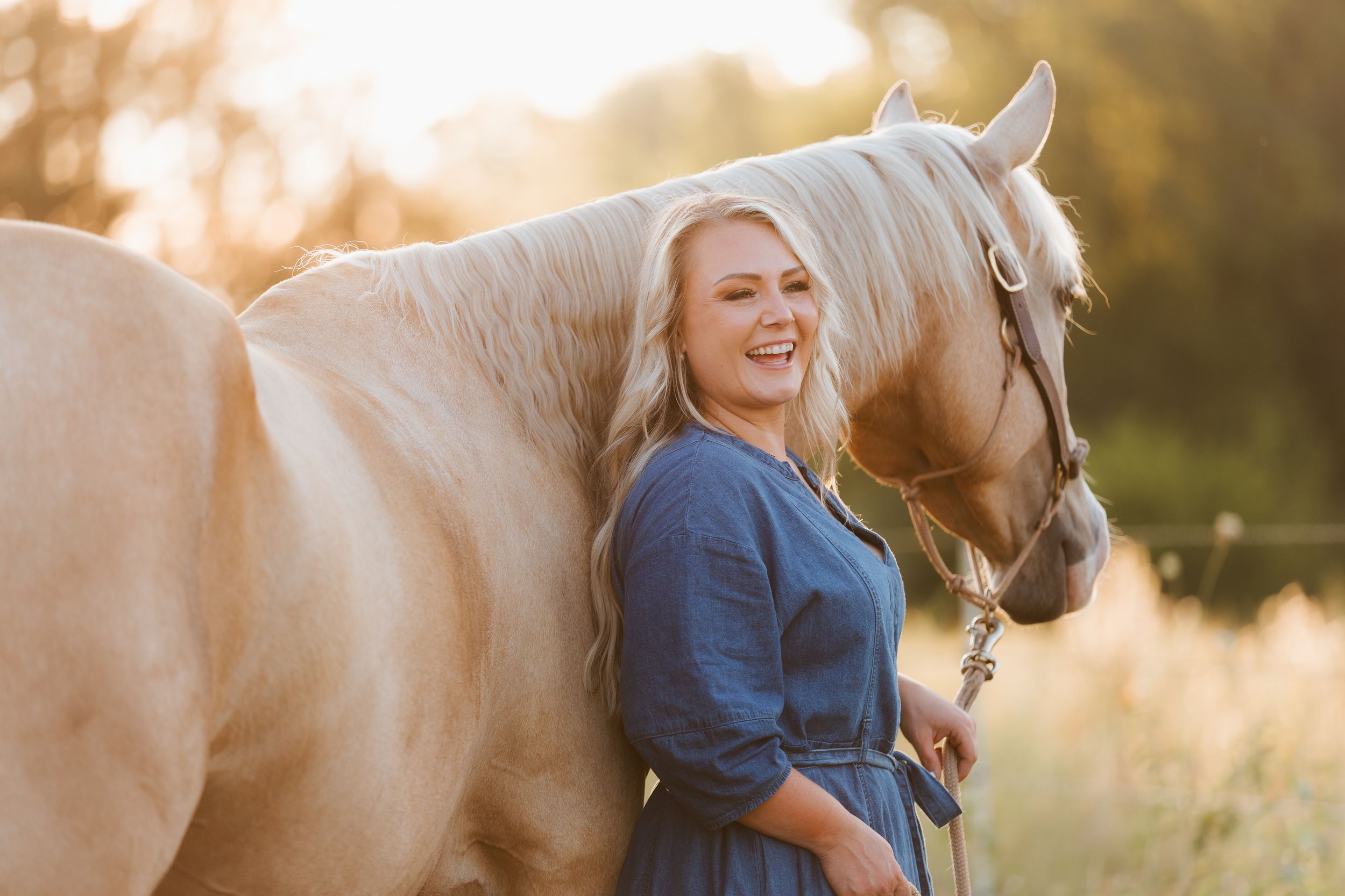Young blonde woman in blue dress smiling beside a palomino horse in a sunlit field.