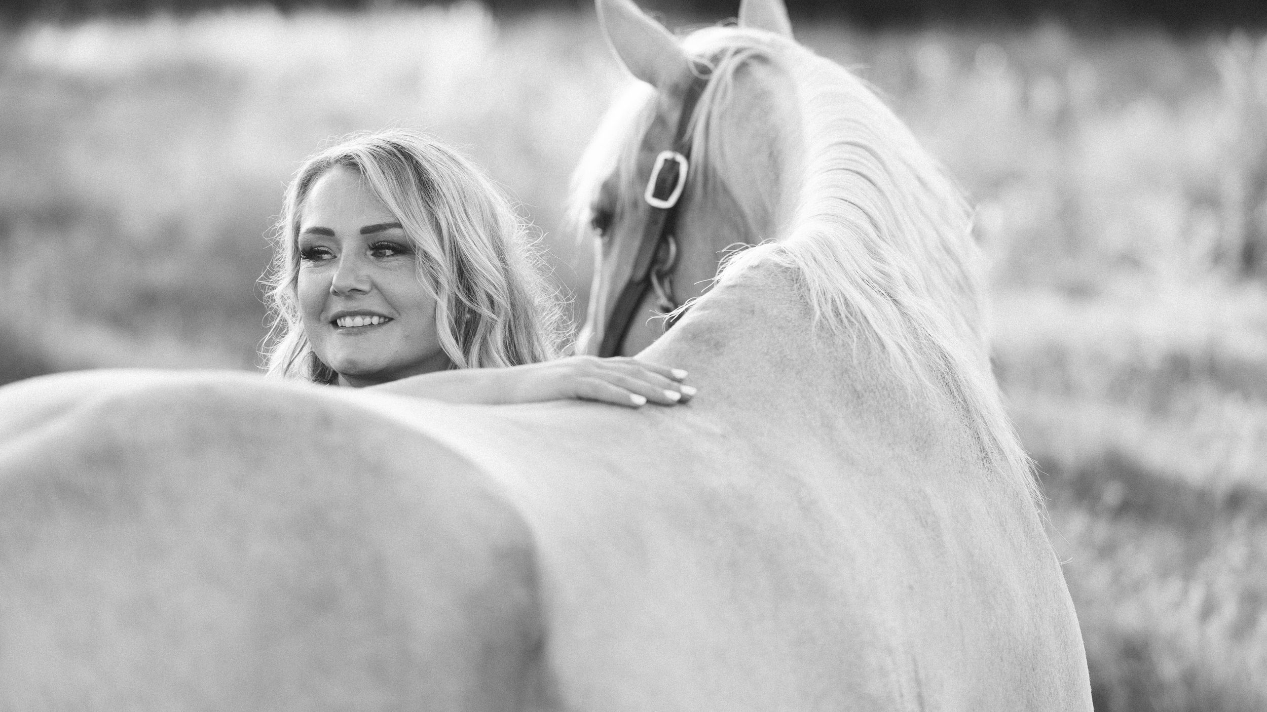 Black and white photo of a smiling blonde woman leaning affectionately against a white horse in a field.
