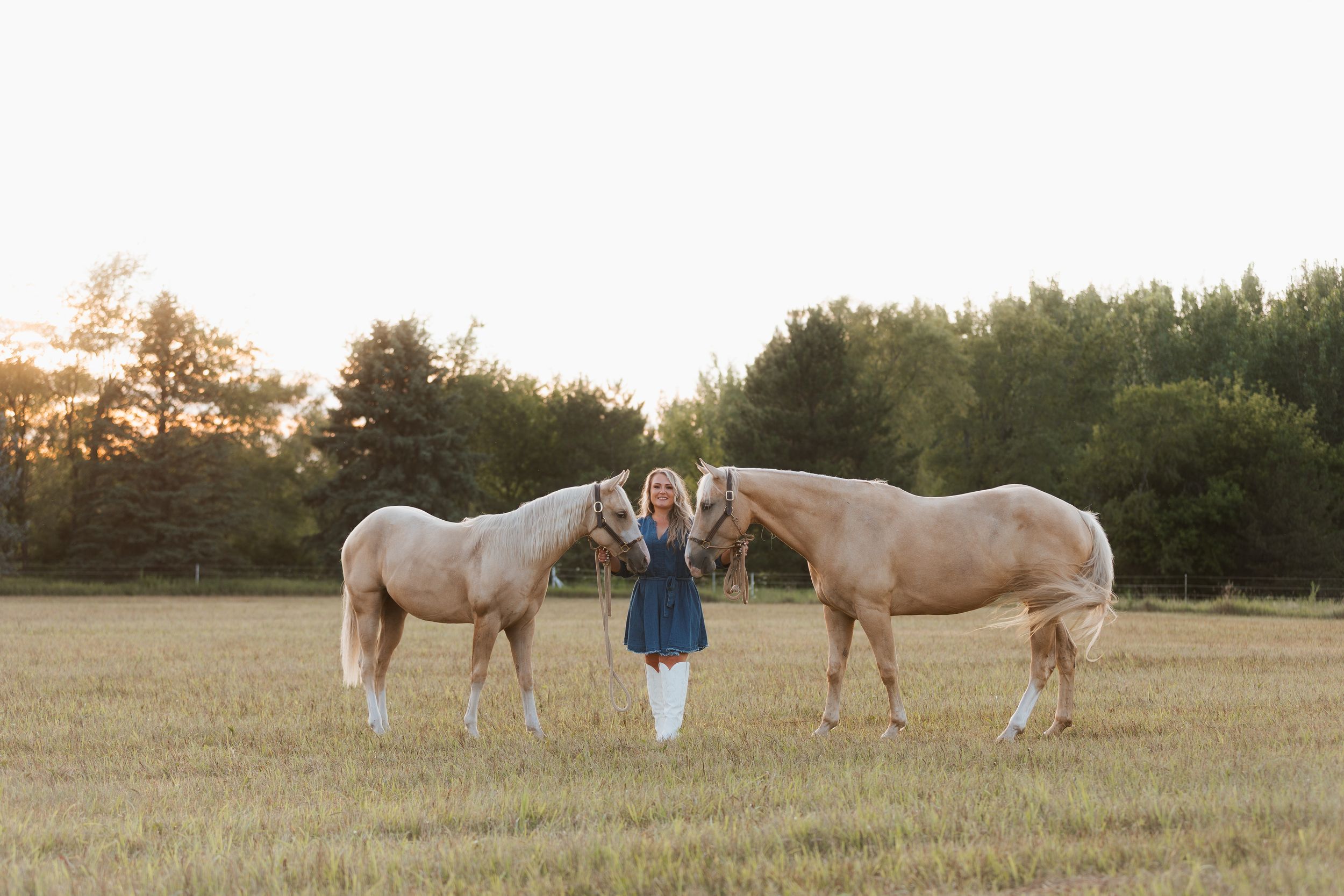 Woman standing between two palomino horses in a sunlit field with trees in the background.