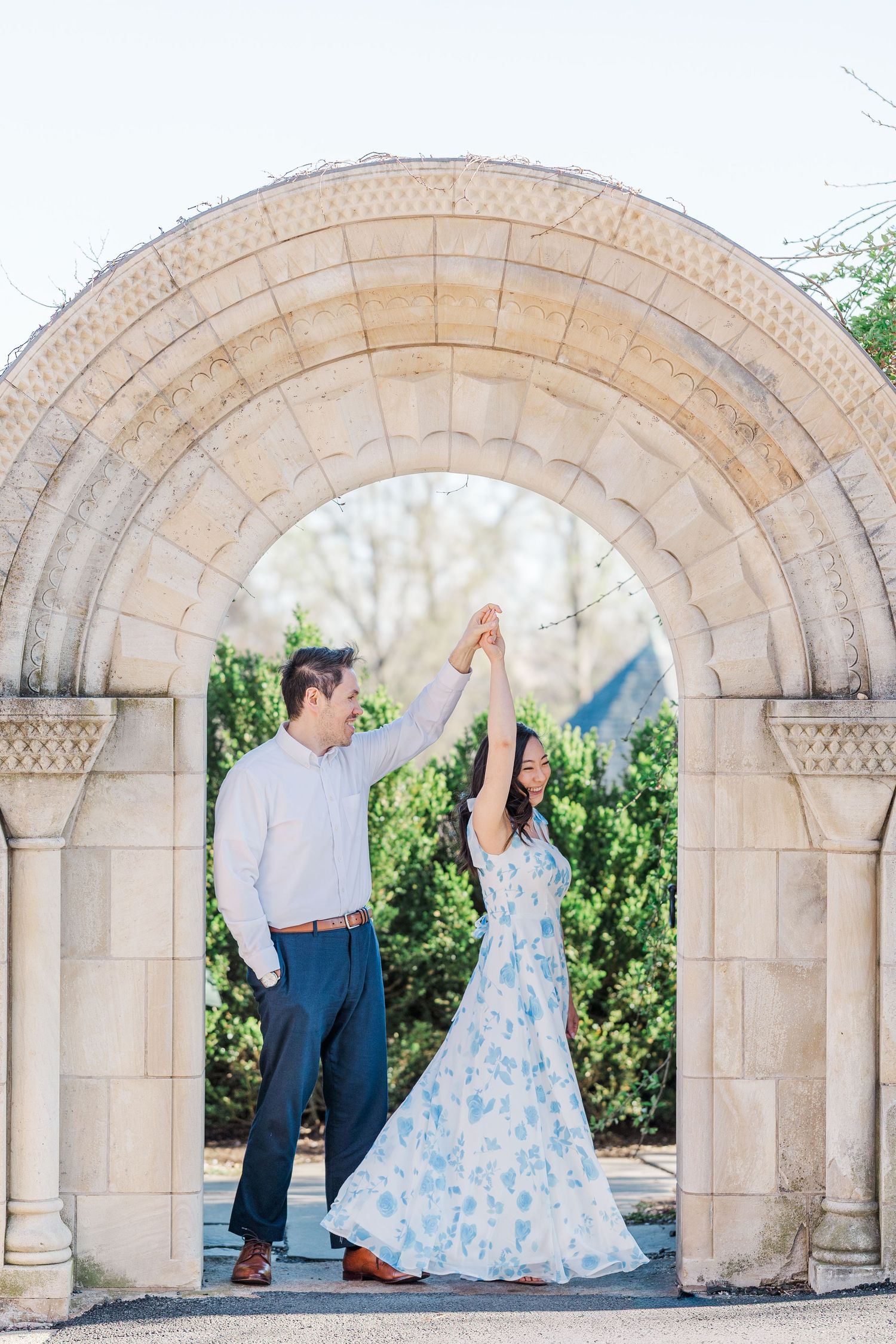 Couple dancing under a stone arch in a garden, woman in floral dress, man in white shirt, lush greenery background.