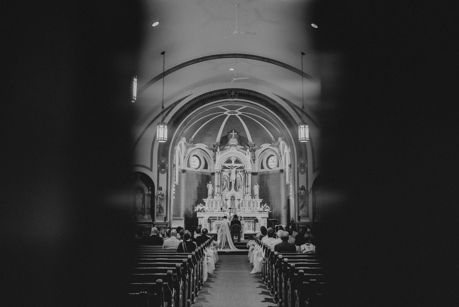 Black and white photo of a wedding ceremony inside a beautiful church with ornate altar and pews filled with guests.