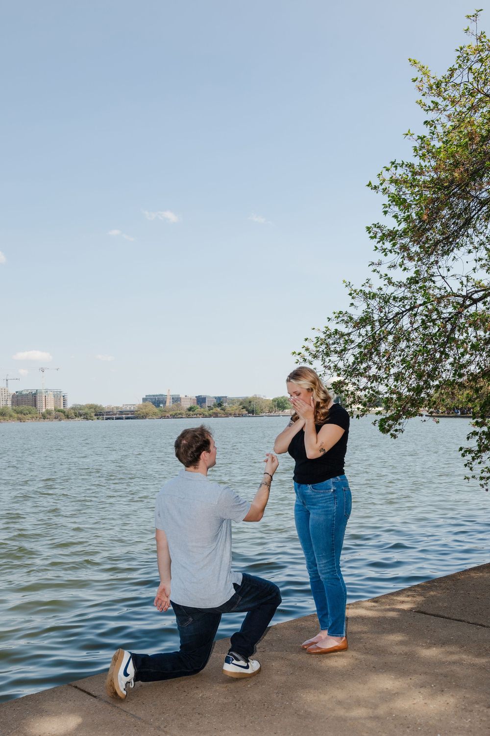 Man kneeling on one knee proposing to woman by a waterfront on a sunny day, with city skyline in background.