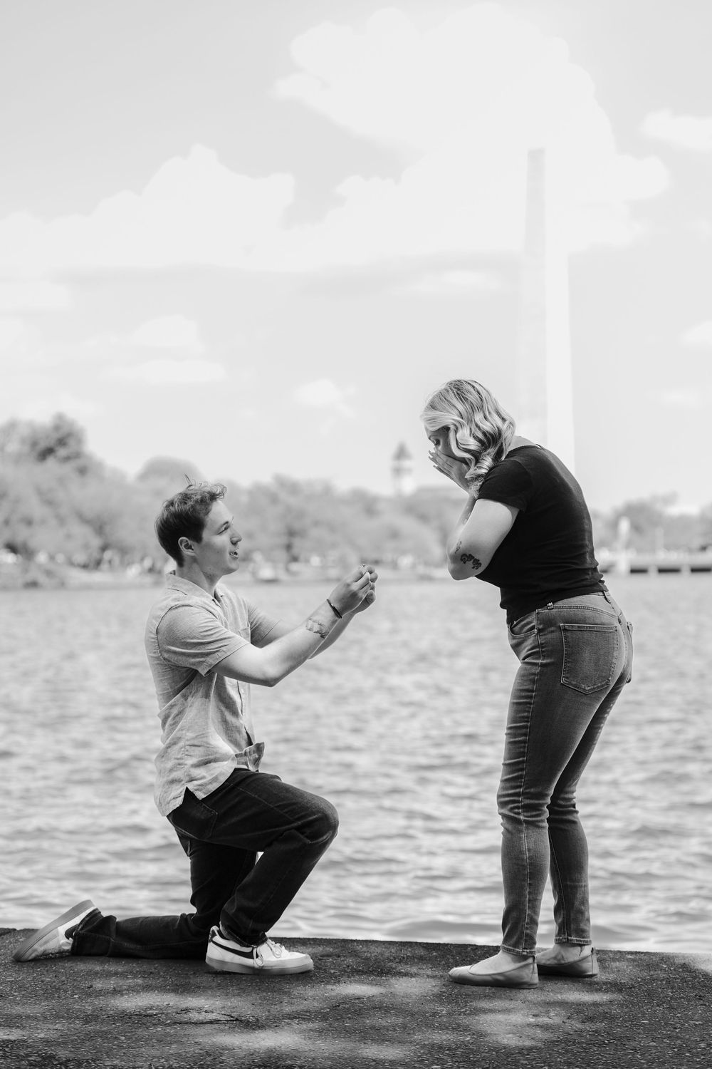 Man proposes on one knee to woman by the Washington Monument reflecting pool in black and white photo.