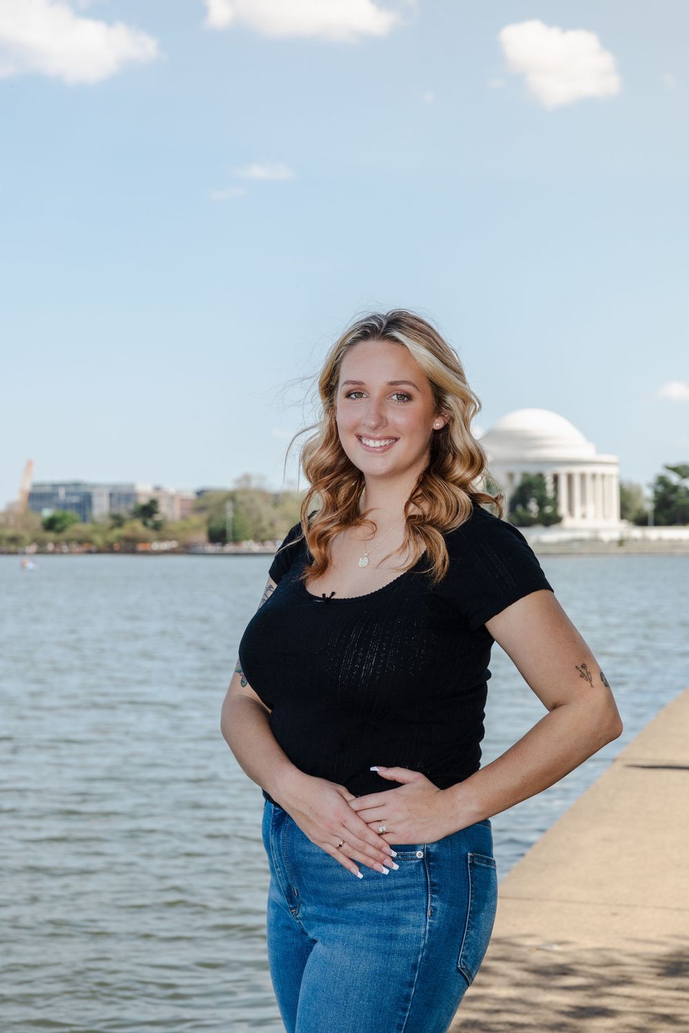 Smiling woman in black top and jeans posing by the Tidal Basin with Jefferson Memorial in background.