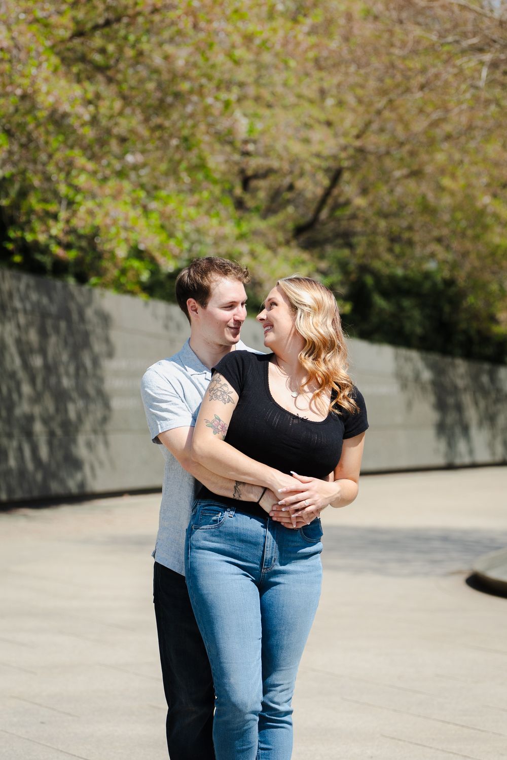 A smiling couple embraces outdoors, the man hugging the woman from behind, both wearing jeans and casual tops with green trees behind.