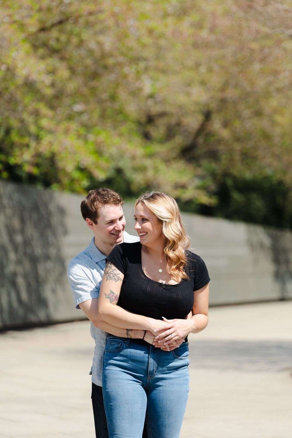 A happy couple shares a tender moment outdoors, man embracing woman from behind, both in jeans with lush green trees in background.