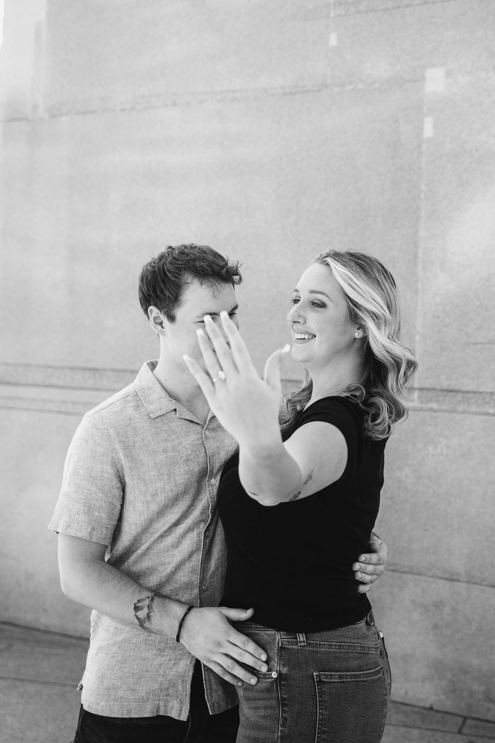 Black and white photo of smiling woman showing engagement ring while man covers his face with hand.
