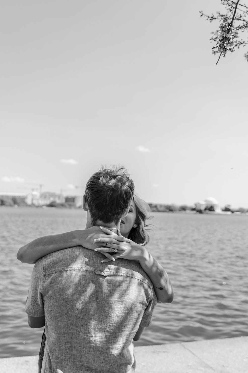 Black and white photo of a couple embracing by a waterfront, woman resting her head on man's shoulder.