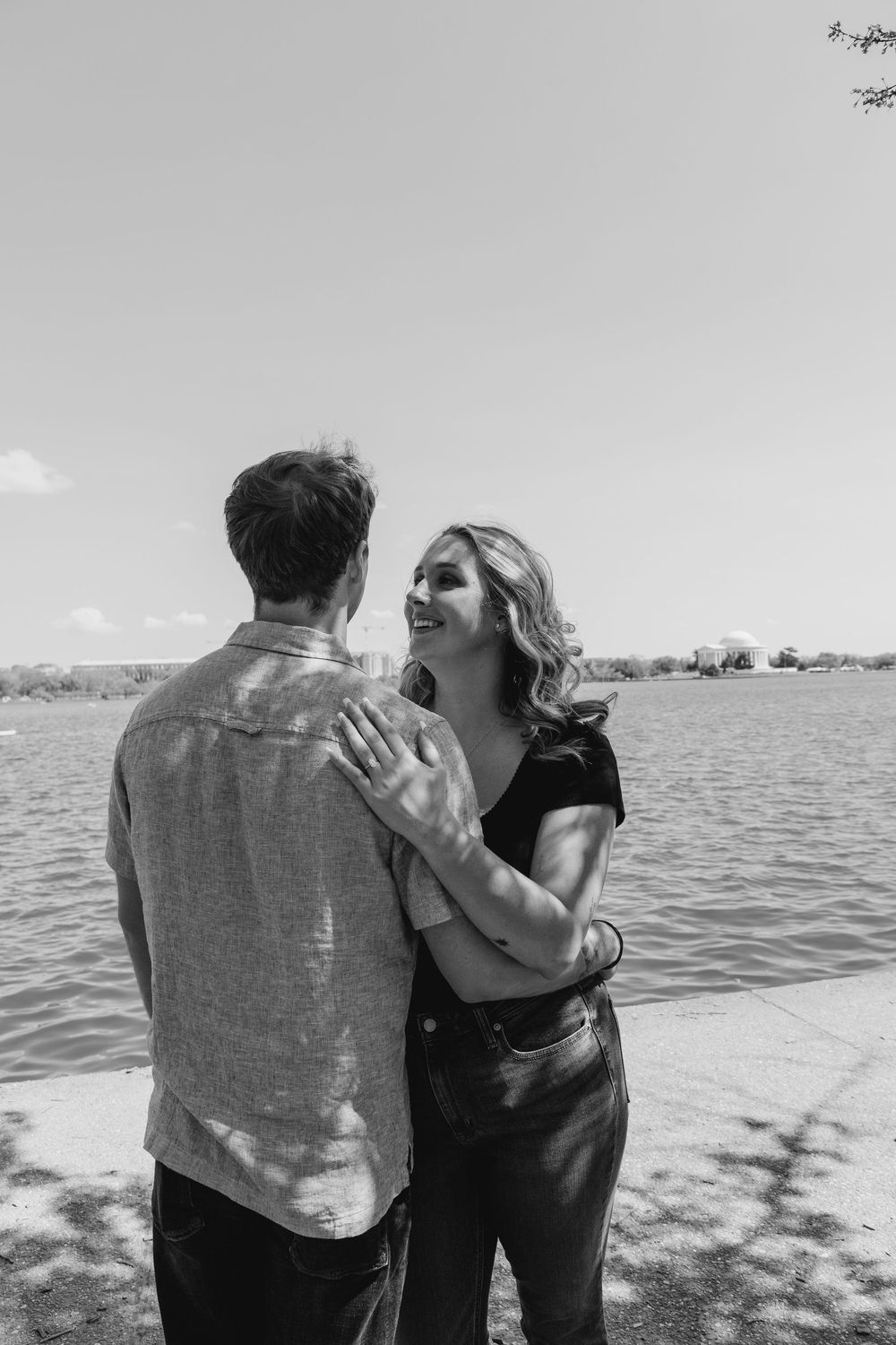 Black and white photo of a smiling woman embracing a man near a lake on a sunny day.