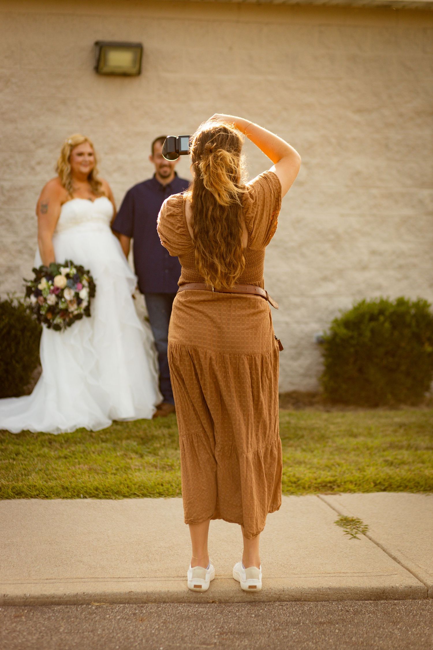 Female photographer in brown dress captures wedding couple posing outdoors against building wall.