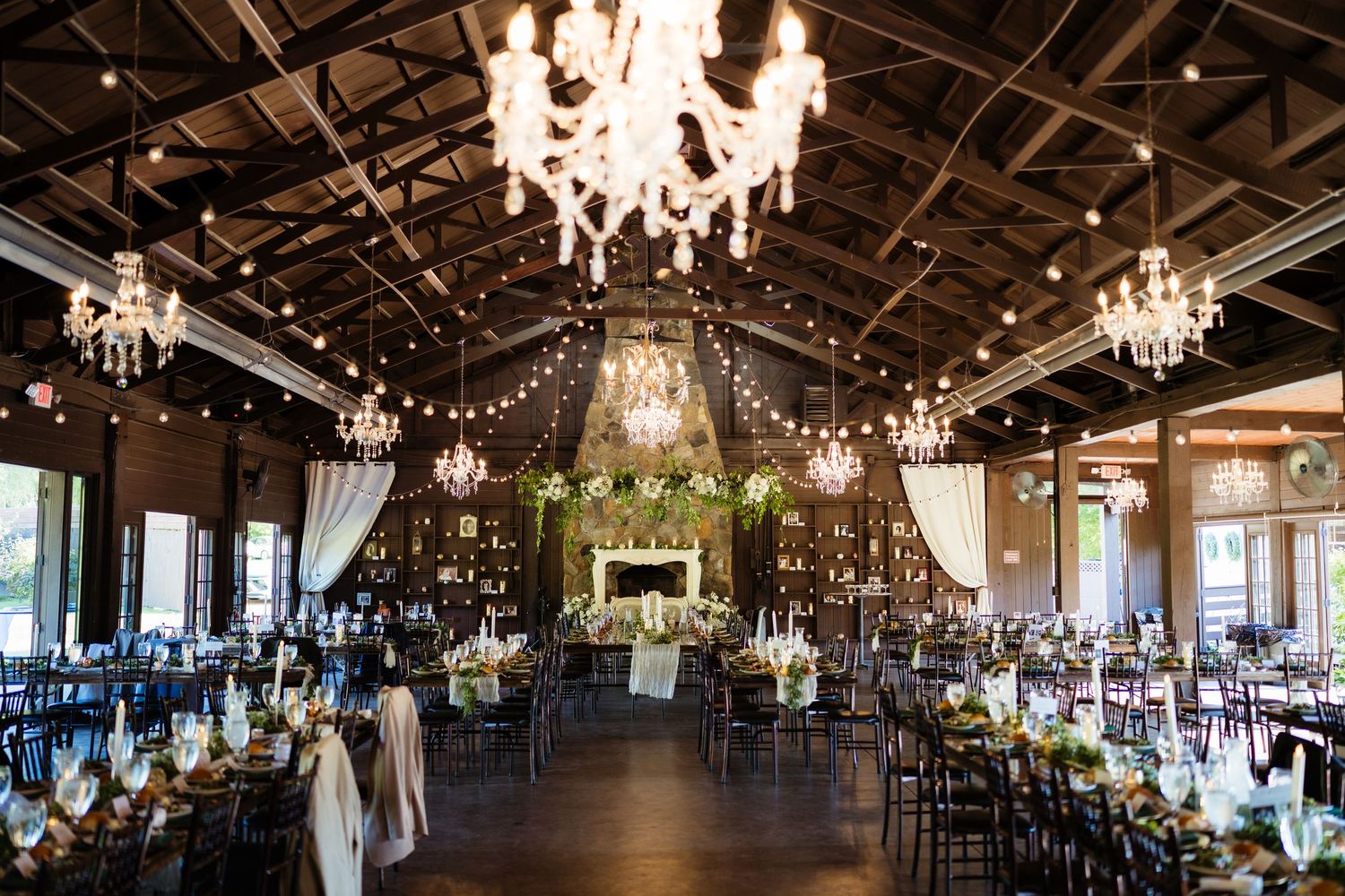 Rustic barn wedding reception venue with string lights, crystal chandeliers, stone fireplace, and elegantly decorated dining tables.