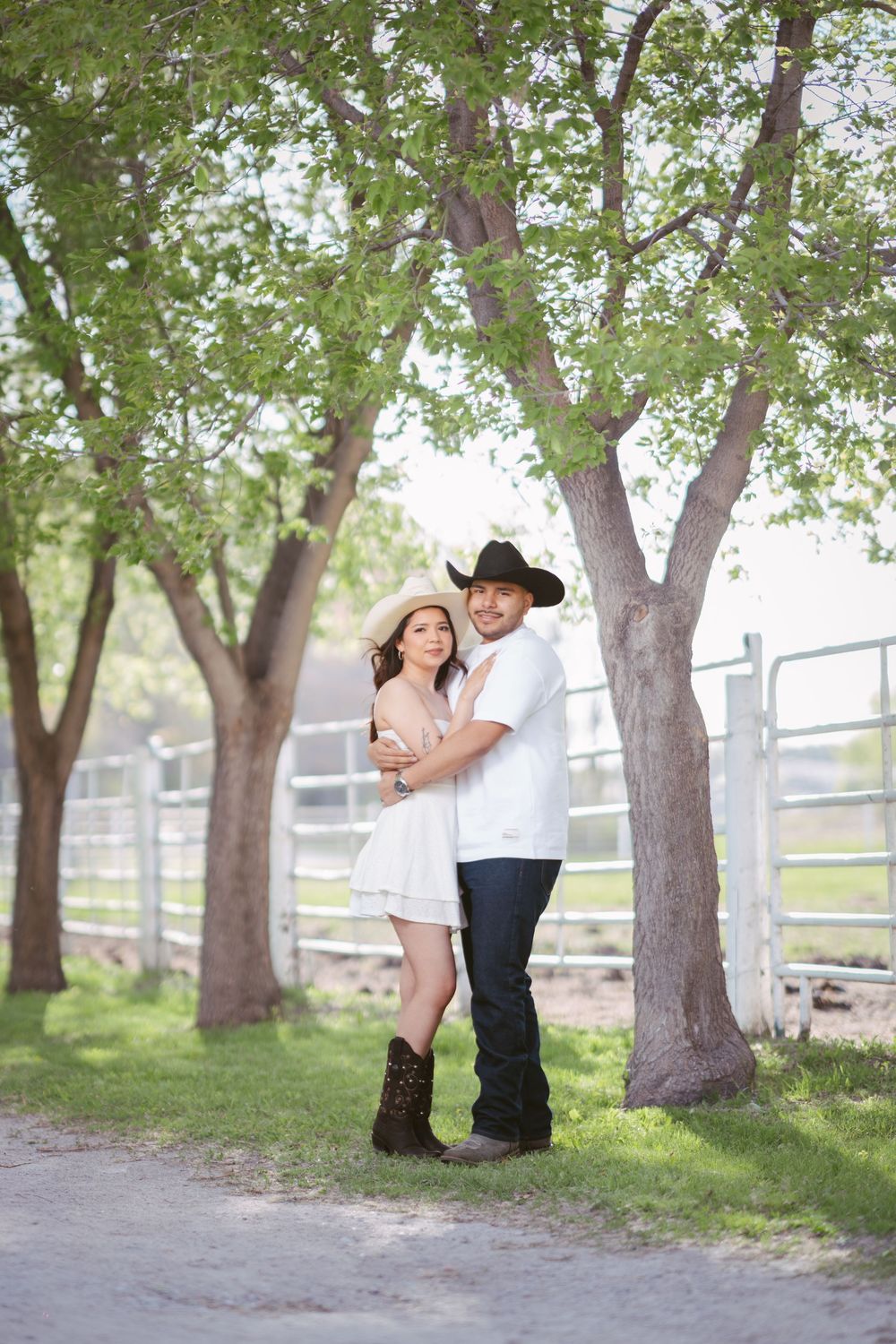 A couple in cowboy hats embraces under green trees near a ranch fence, wearing white outfits and cowboy boots.